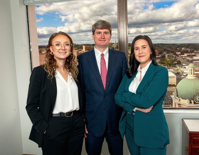 Three professionally dressed people posing in front of a city view through large windows