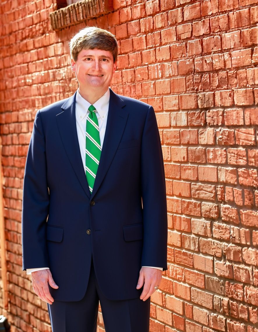 Man in a navy suit and green striped tie standing beside a brick wall outdoors