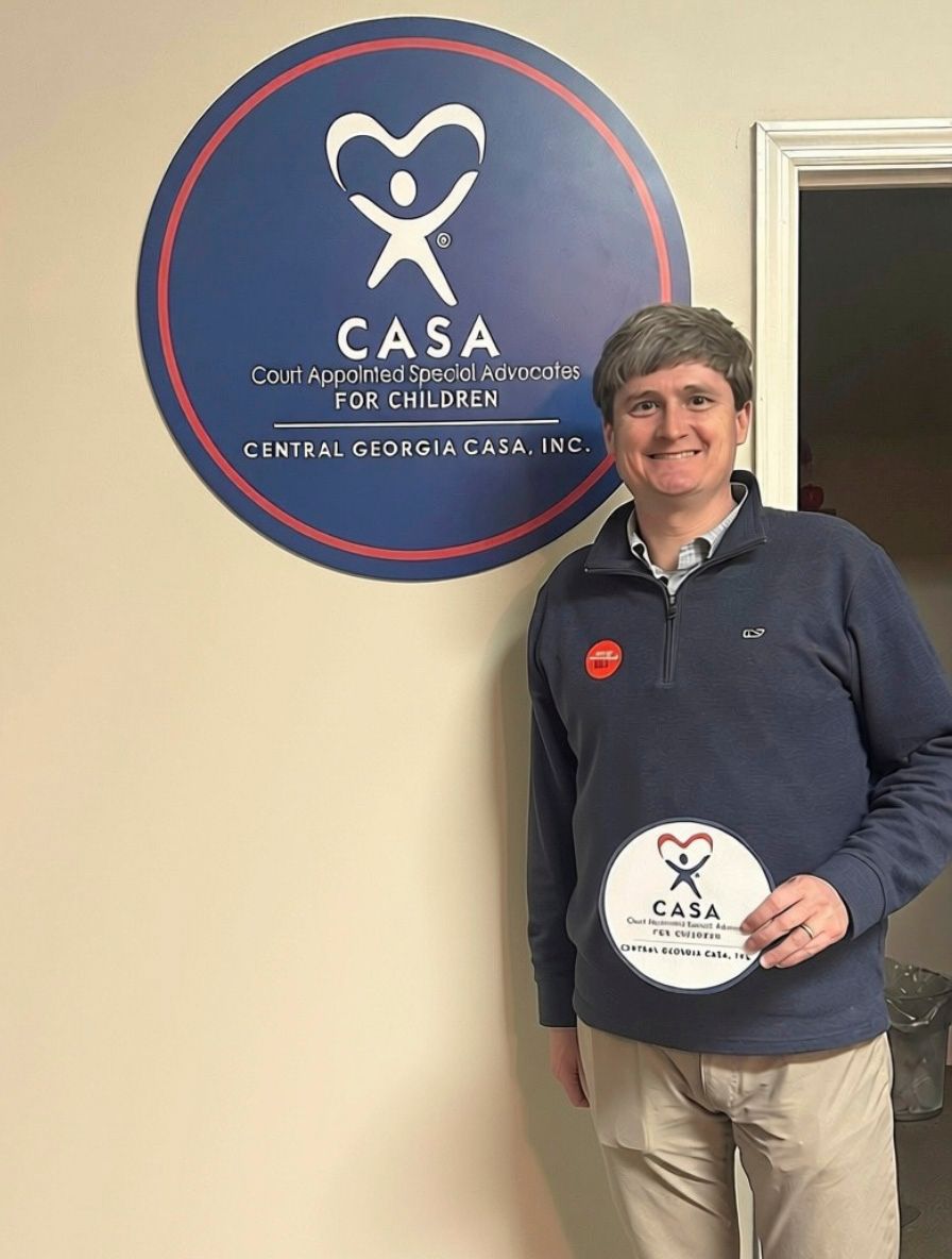 Person standing indoors beside a CASA sign, holding a round CASA plaque and smiling.