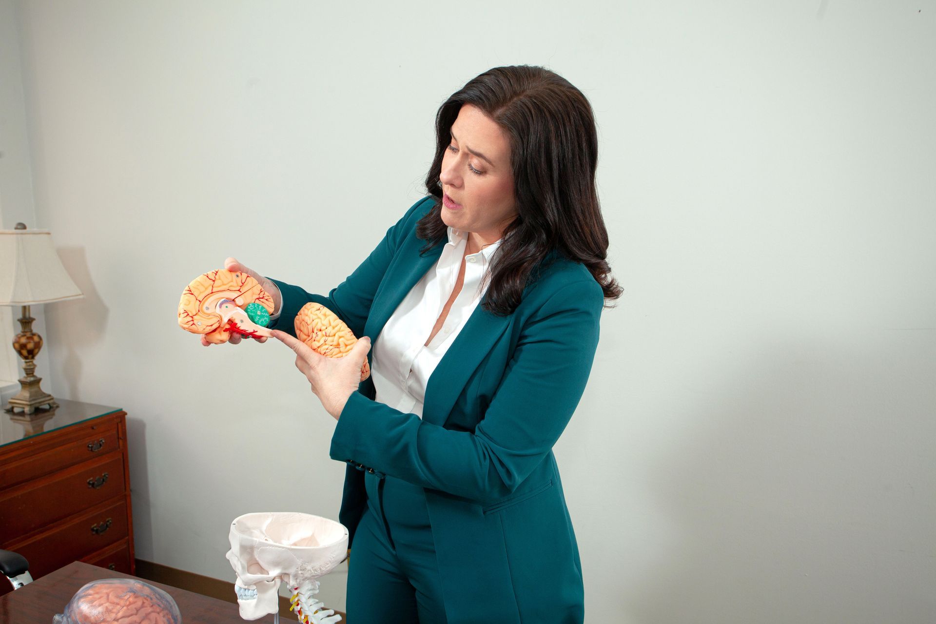 Woman in teal blazer examines an orange brain model beside a tissue box in an office.