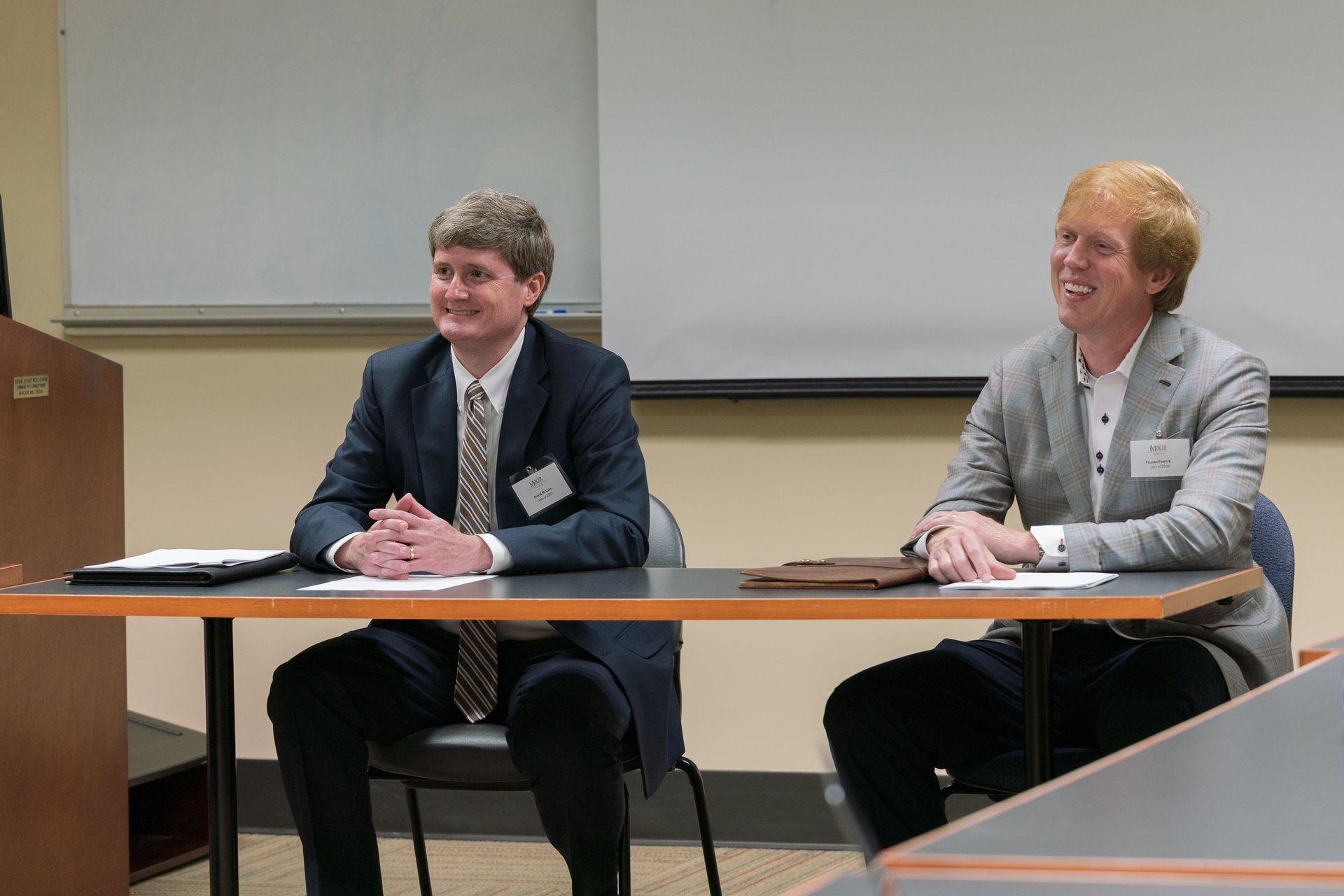 Two people seated at a table in a classroom, facing forward during a presentation.