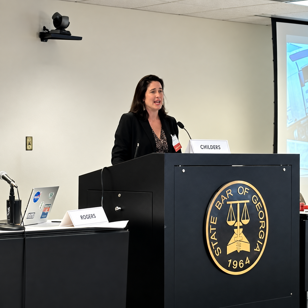 Speaker at a podium in a classroom, presenting beside a projected screen.