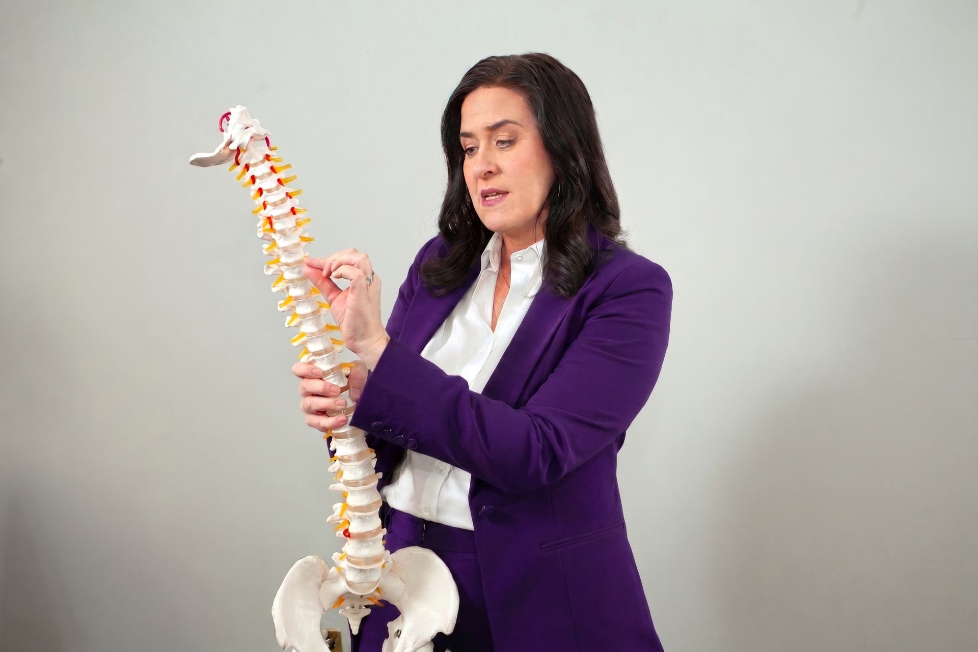 Woman in purple blazer demonstrating a human spine model against a plain background