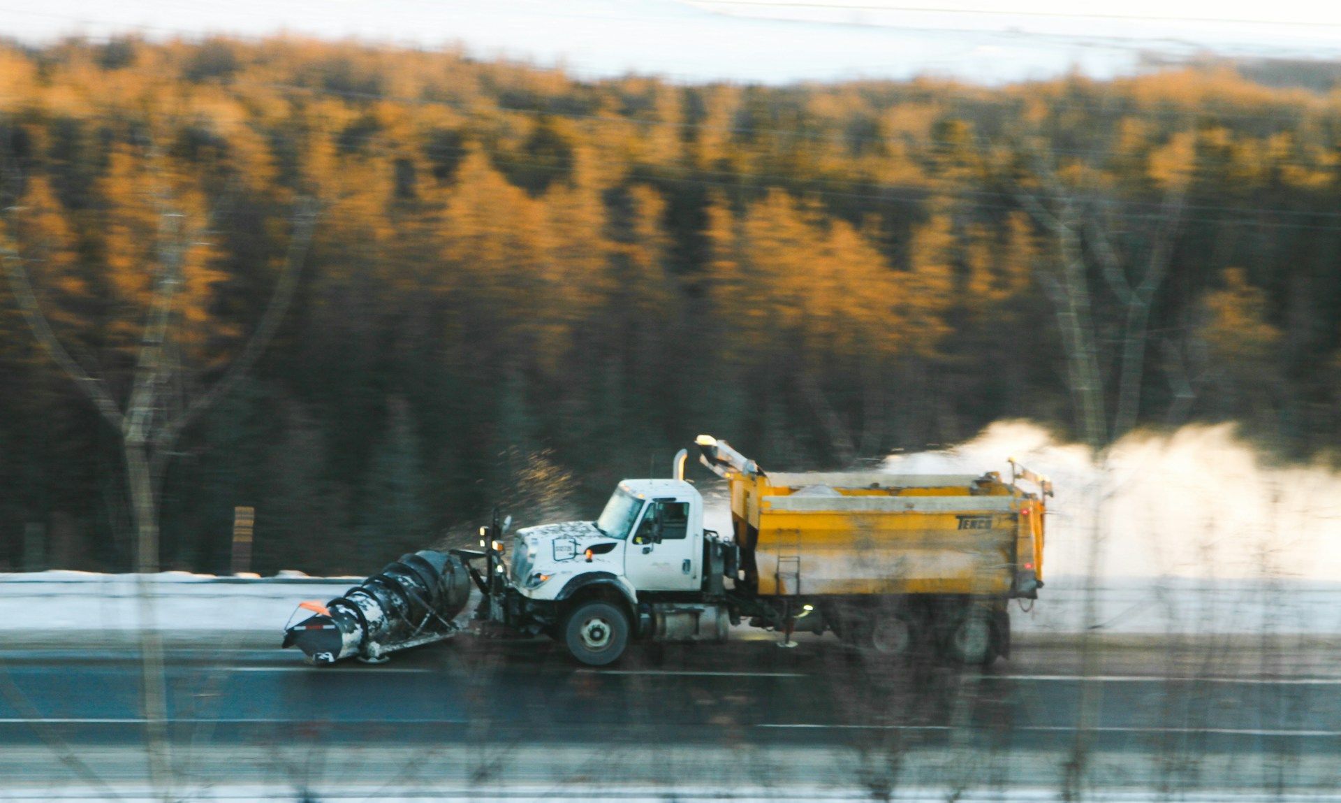 Snowplow clearing a wet road with a forest of autumn trees in the background
