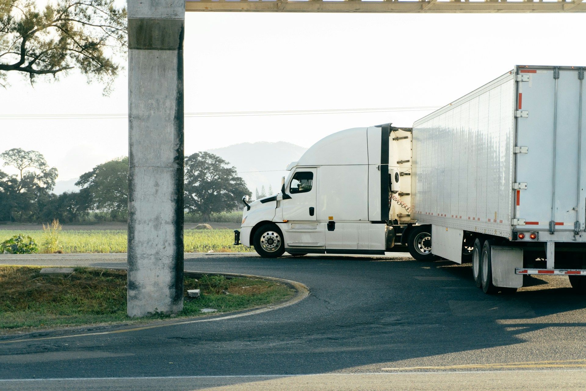 White semi-truck pulling a box trailer around a curve on a rural road near a concrete pillar