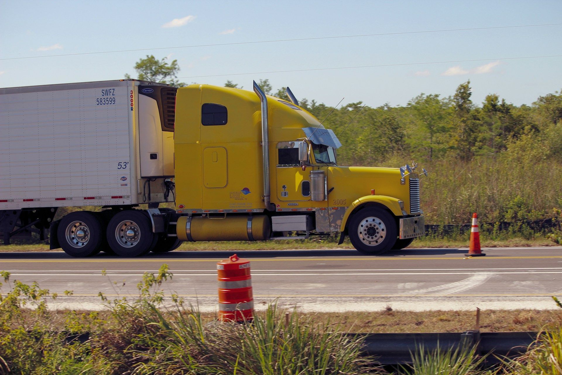 Yellow semi-truck on a highway shoulder beside an orange traffic cone, with trees in the background