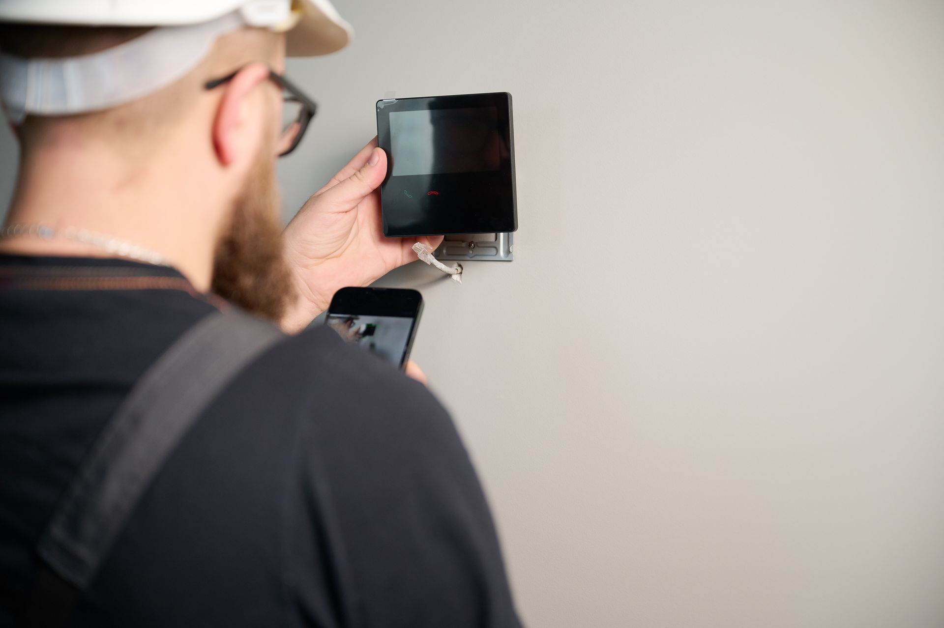 Person using a smartphone to inspect a wall-mounted black device while holding a brush or roller Person using a smartphone to inspect a wall-mounted black device while holding a brush or roller