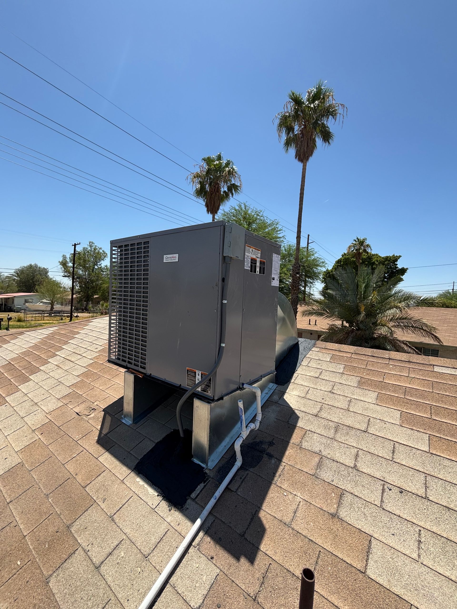 Gray HVAC unit on a brown-shingled roof under a clear, blue sky with palm trees in the background.