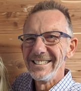 Smiling man wearing glasses and a checkered shirt, standing against a wooden wall.