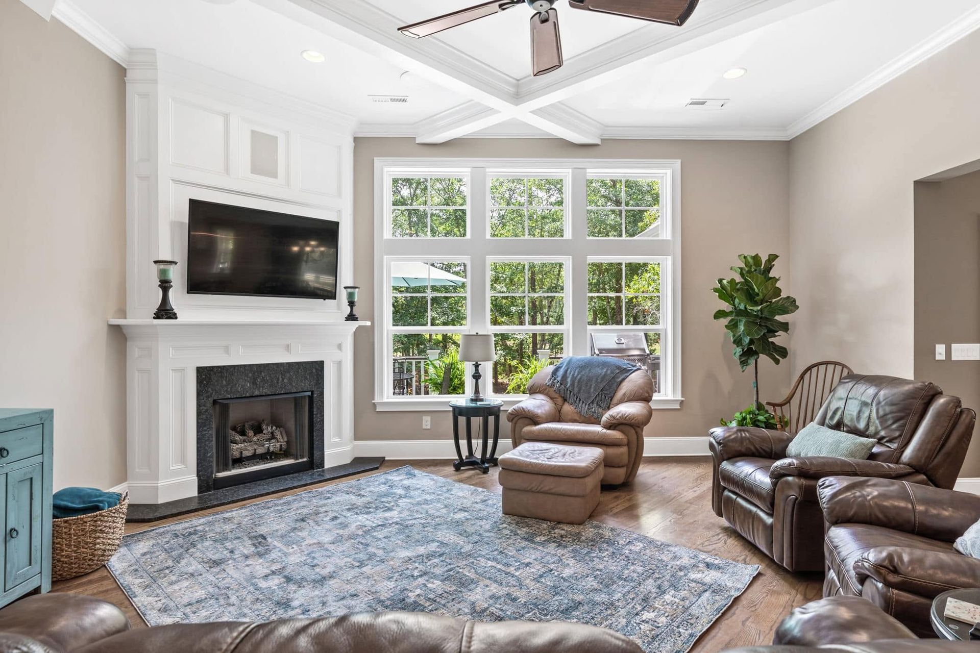Bright living room with fireplace, large windows, leather chairs, blue rug, and ceiling fan