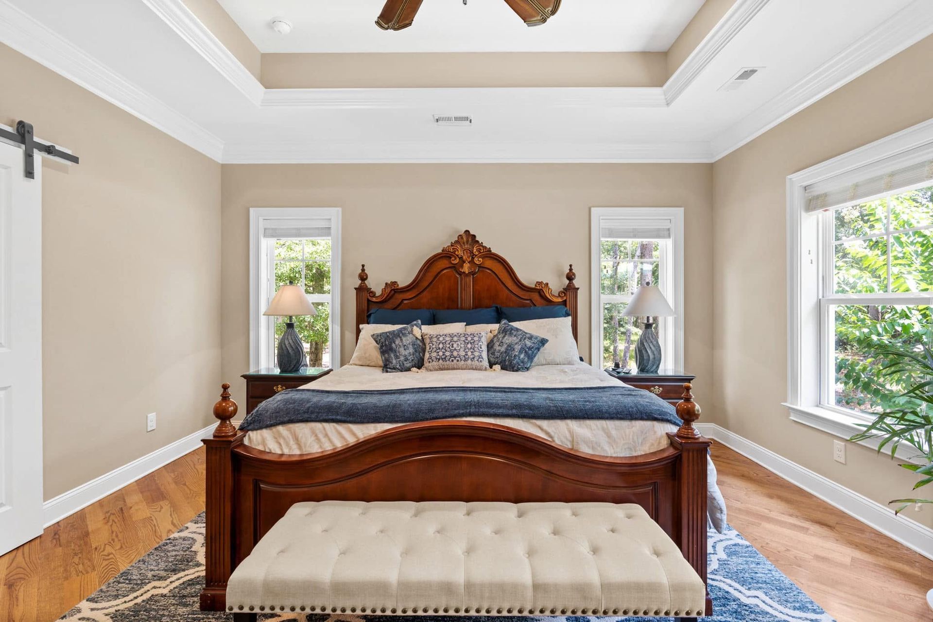 Bright bedroom with a carved wooden bed, tufted bench, blue rug, and large windows