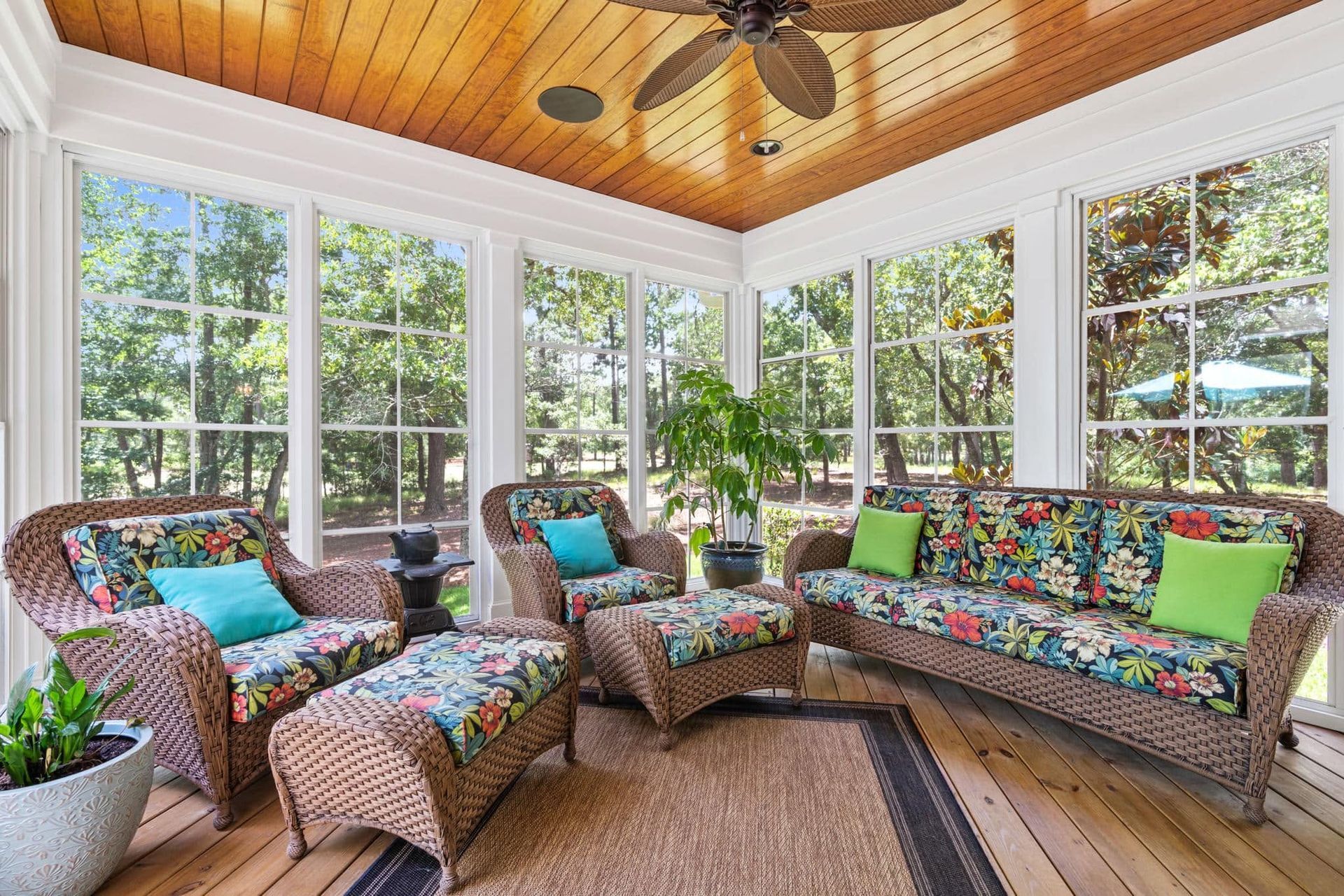 Bright sunroom with wicker seating, colorful cushions, potted plants, and large windows under a wood ceiling.