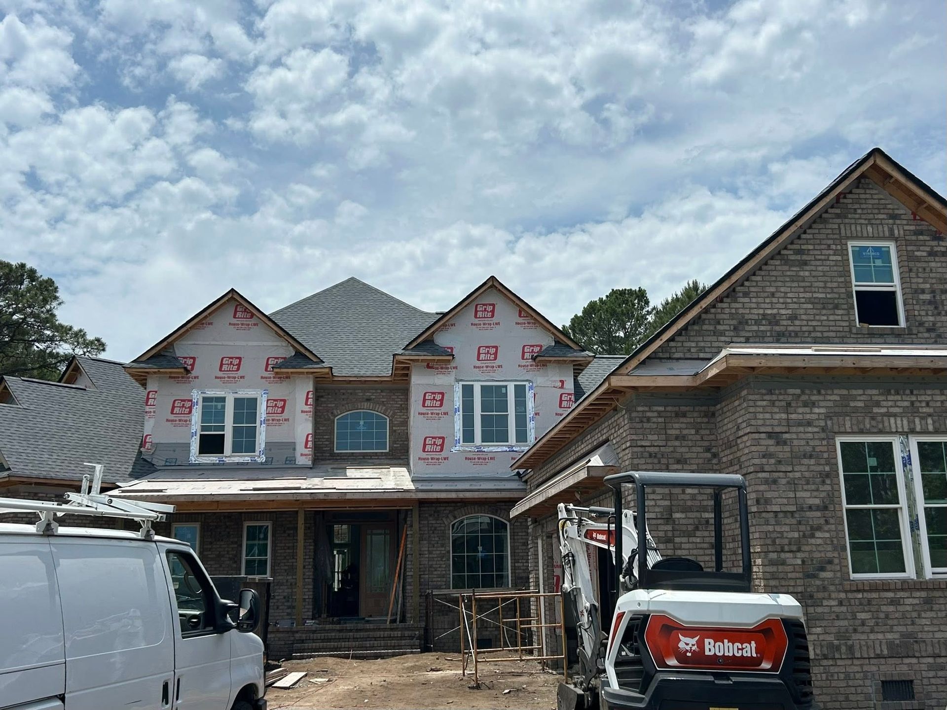 Suburban house under construction with gray siding, pink window trim, a white van, and a Bobcat loader in front