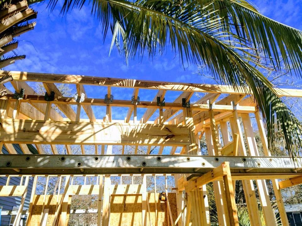Wooden house frame under construction against a bright blue sky with palm fronds overhead