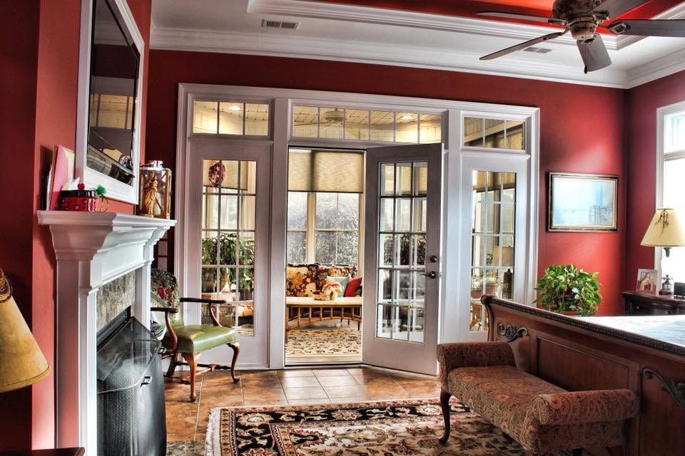 Cozy living room with red walls, fireplace, French doors, and patterned rug opening to a bright sunroom