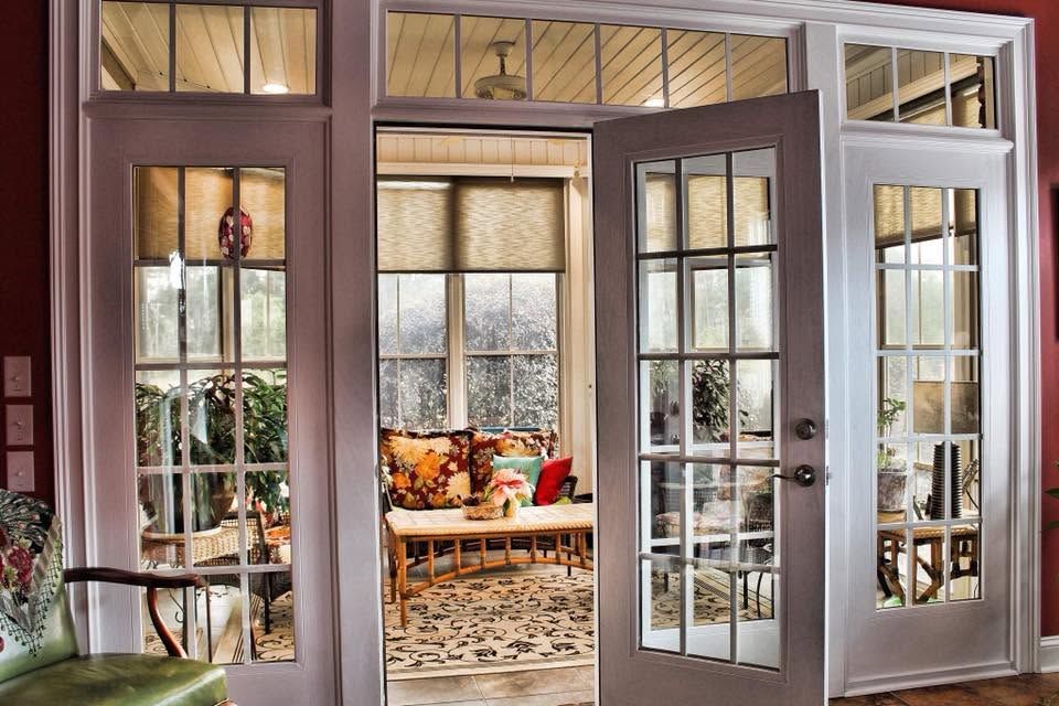 Open French doors to a cozy sunroom with wicker seating and patterned curtains