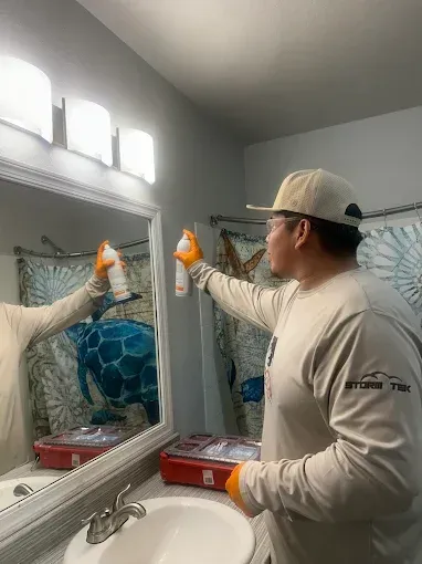 Person painting a bathroom mirror frame white while wearing a cap and gloves.
