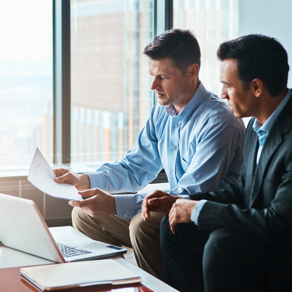 Two men in office setting reviewing documents with a laptop.