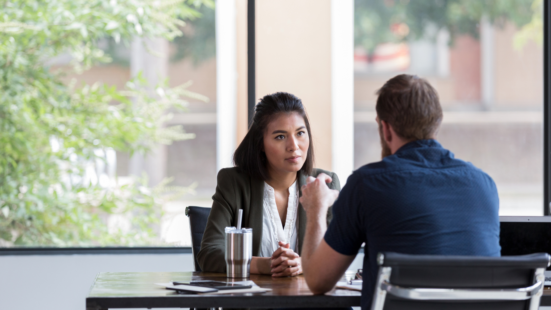 Woman in blazer and man in blue shirt seated at table, conversing indoors.