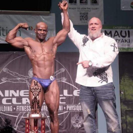 Bodybuilder flexes on stage, holding trophy with arm raised, next to a man pointing.