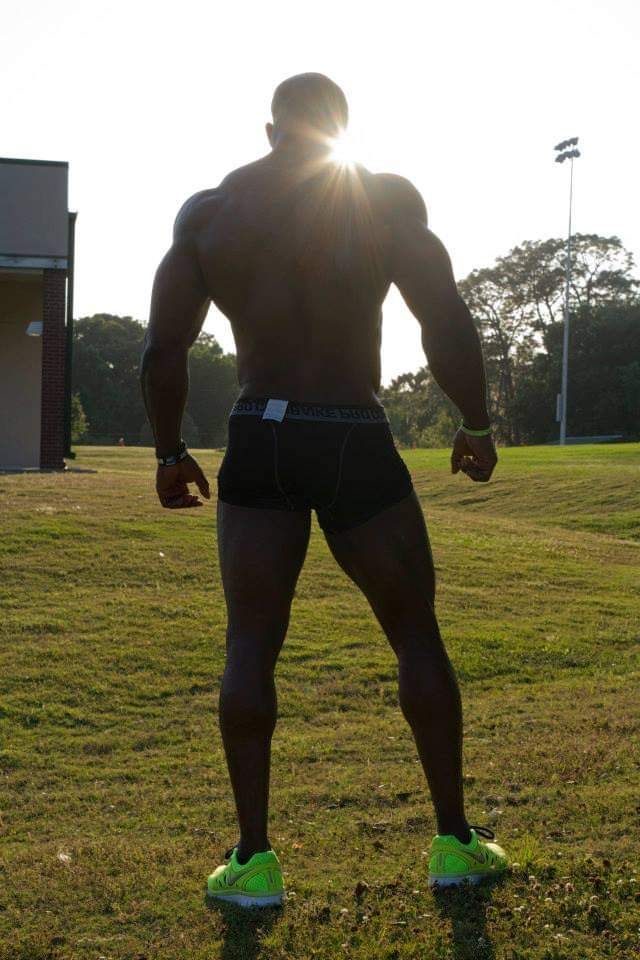 Muscular person in athletic shorts and neon sneakers stands on grass with sun shining behind.