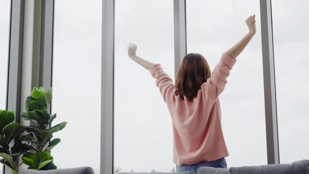 Person in a pink sweater raises both arms while looking out a bright window.