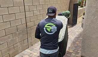 Worker in a black cap and shirt stands in a brick walkway beside a wall, handling equipment.