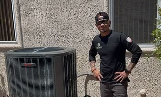 Person standing beside a large outdoor air conditioner unit in front of a house, hands on hips.