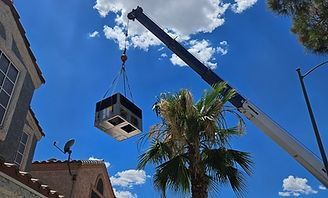 Crane lifting a black cube near palm trees and buildings against a bright blue sky