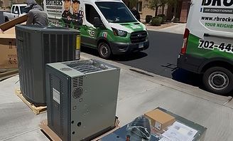 Large gray HVAC units on a city sidewalk beside parked green moving vans and a poster board.