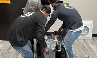 Three workers bending over an open appliance on the floor in a room with wood flooring and a dryer behind them