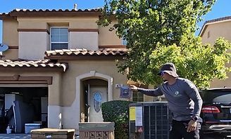 Person standing by an air conditioner outside a beige two-story house, pointing at it in sunlight