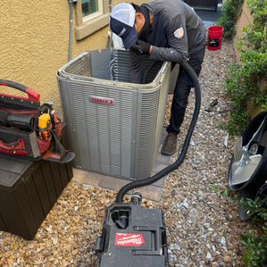 Technician servicing an outdoor HVAC unit, leaning into the open top of the equipment beside a house.
