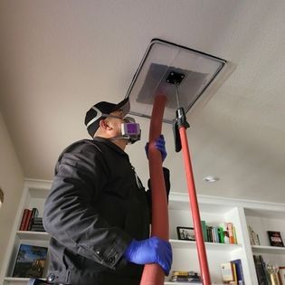 Person cleaning a ceiling vent with a vacuum hose while wearing gloves and a respirator mask