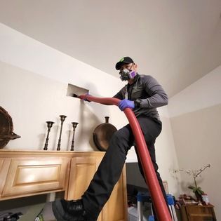 Person using a vacuum cleaner on top of a cabinet in a living room, with a red hose and cleaning tools nearby
