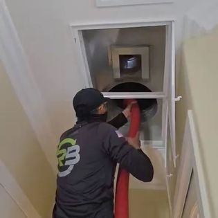 Worker in black uniform uses a red hose to clean a wall vent opening indoors.