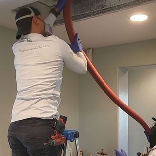 Worker using a red vacuum hose to clean debris from a ceiling vent in a room