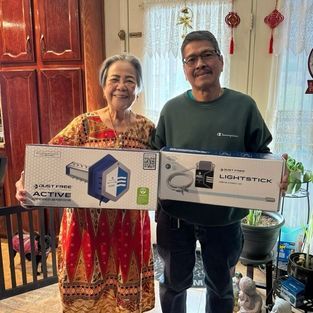 Smiling couple holding boxed lights indoors, with hanging decorations in the background