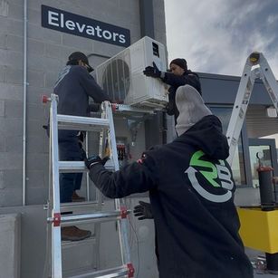 Workers installing an air conditioner beside an “Elevators” sign, with a ladder in a building hallway