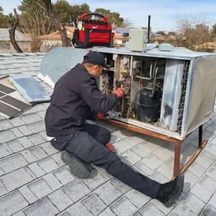 Technician servicing rooftop HVAC unit beside a red tool bag on a shingled roof