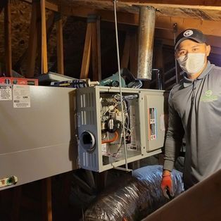 Technician standing beside an open HVAC furnace in an attic crawlspace, wearing a mask and gloves.