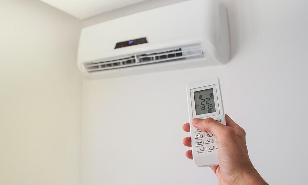 Hand holding a remote toward a wall-mounted air conditioner with the display on, in a white room
