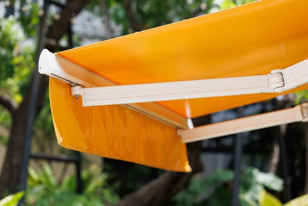 Orange retractable awning with white support arms against a leafy outdoor background — Tully Blinds in Tully, QLD