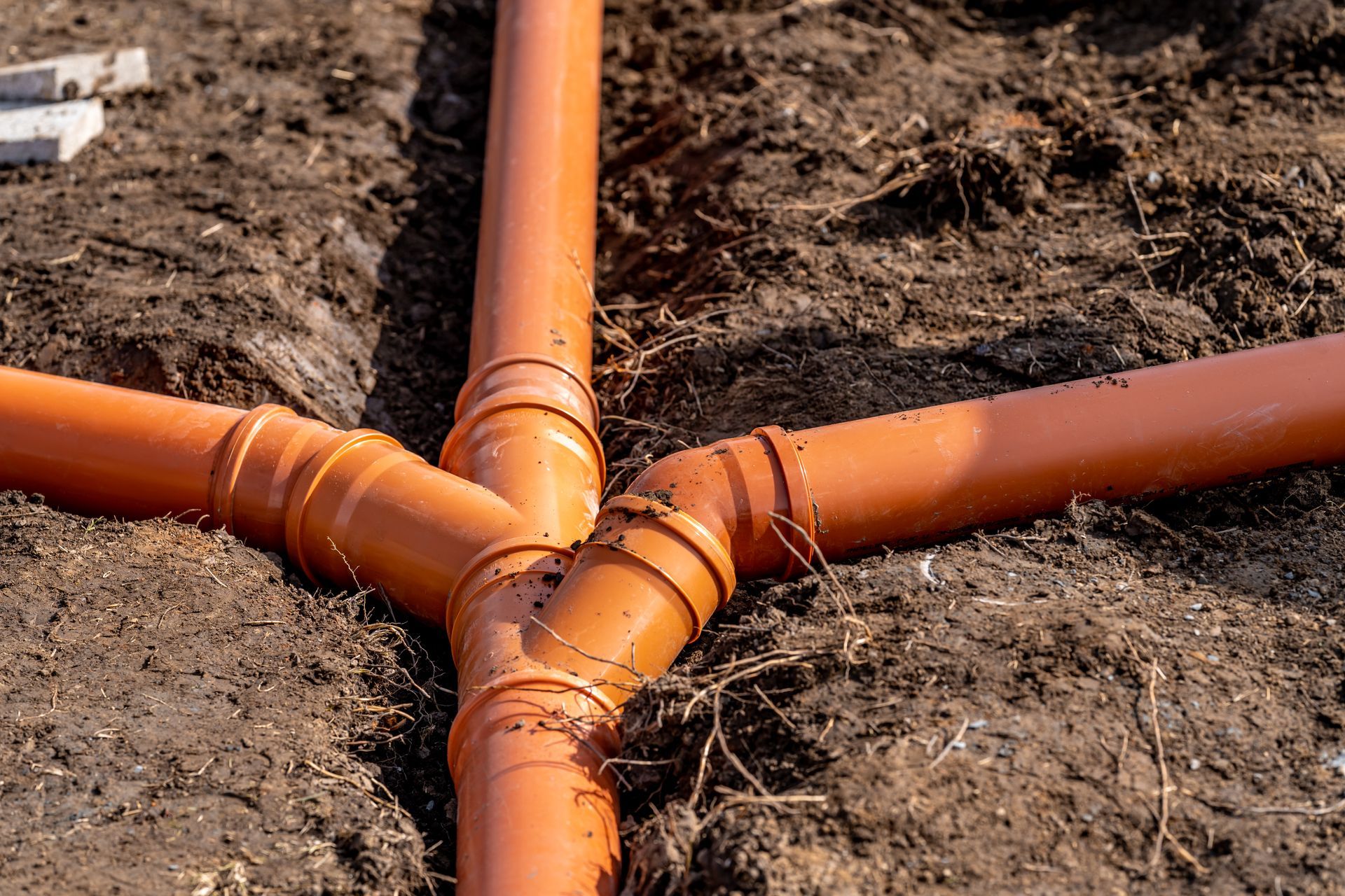 An orange PVC Y-junction pipe connected to three pipe segments embedded in a dirt trench.