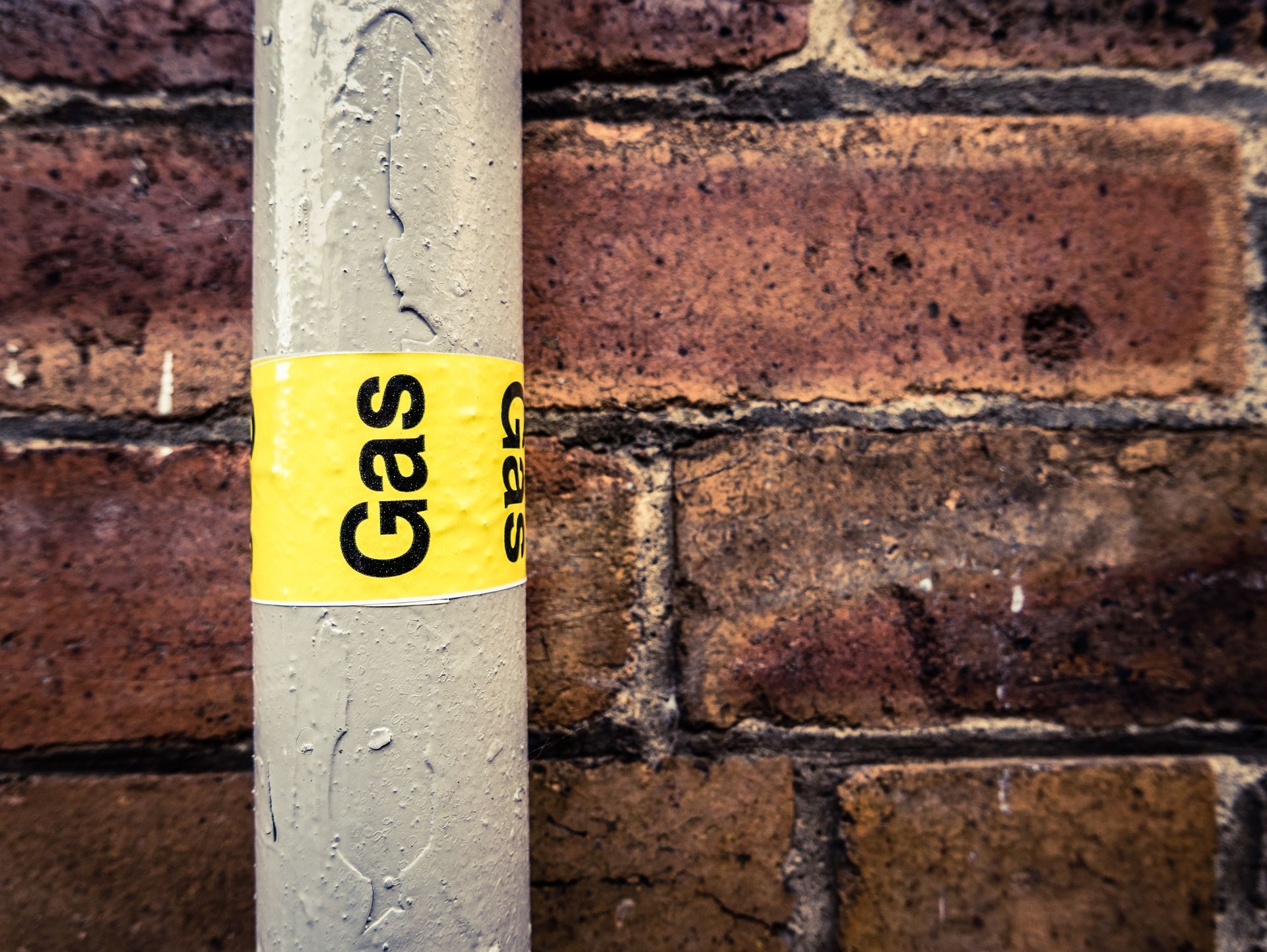 A grey metal pipe against a brick wall, featuring a yellow warning label with the word