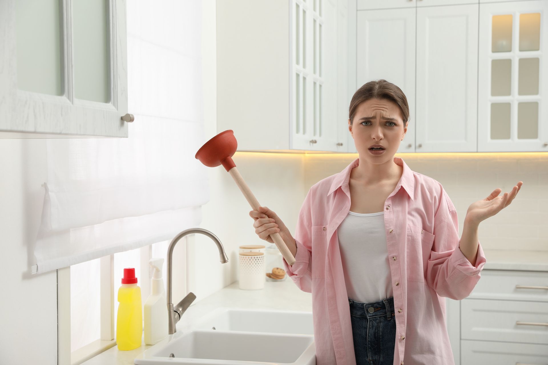 A person in a pink shirt looks frustrated while holding a plunger in a white kitchen with a sink.