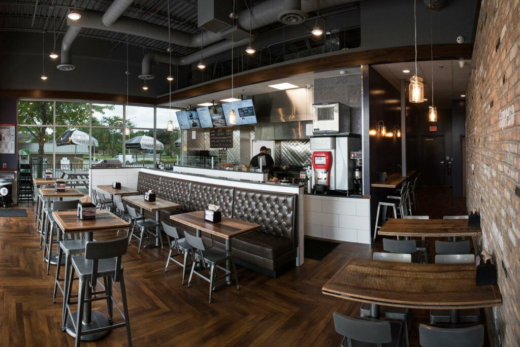 Restaurant interior: wooden tables, brick wall, open kitchen, bar seating, large window, dark gray ceiling.