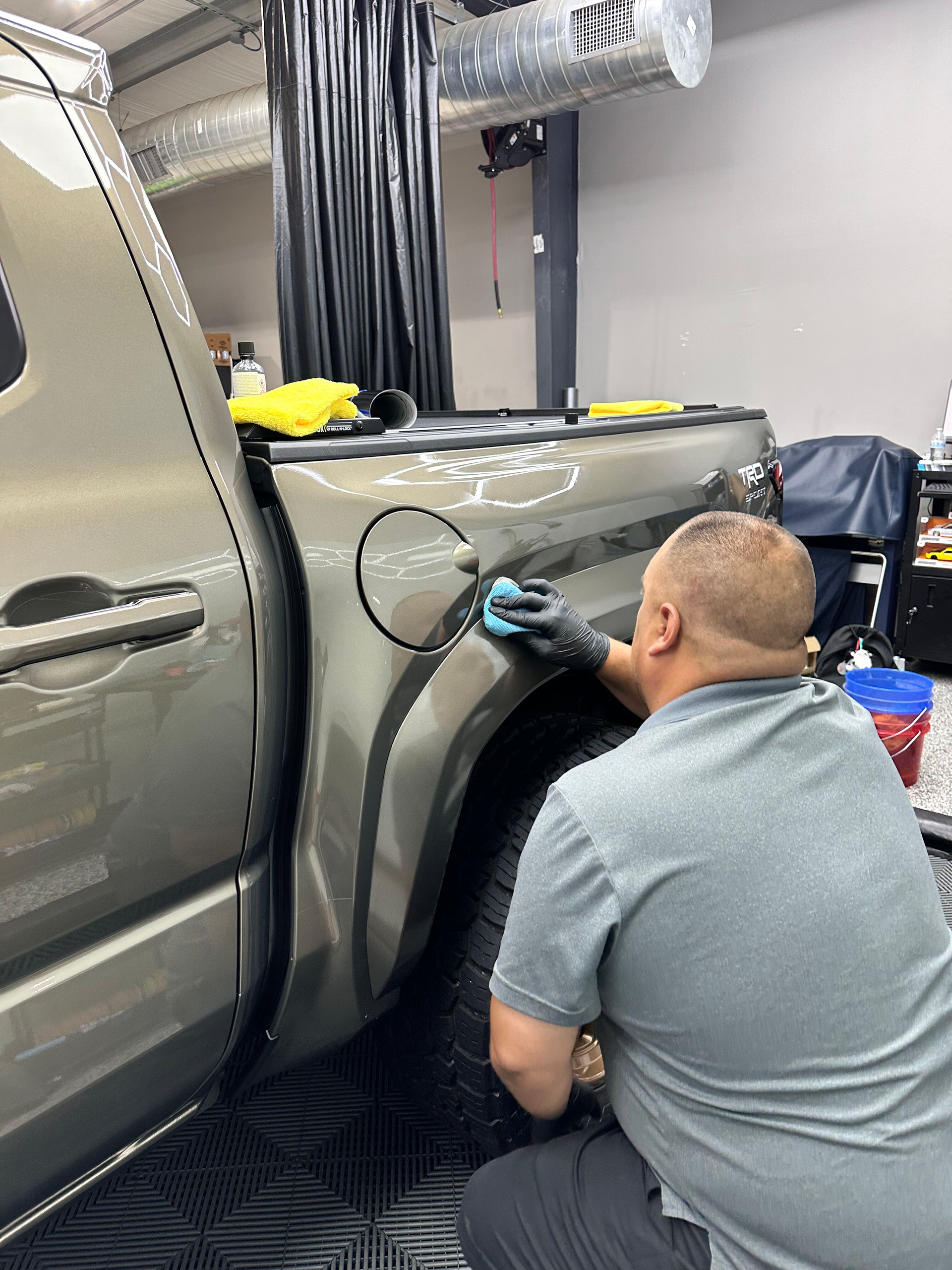 A man in a gray shirt and gloves polishes a gray pickup truck's fender near the gas tank in a garage.