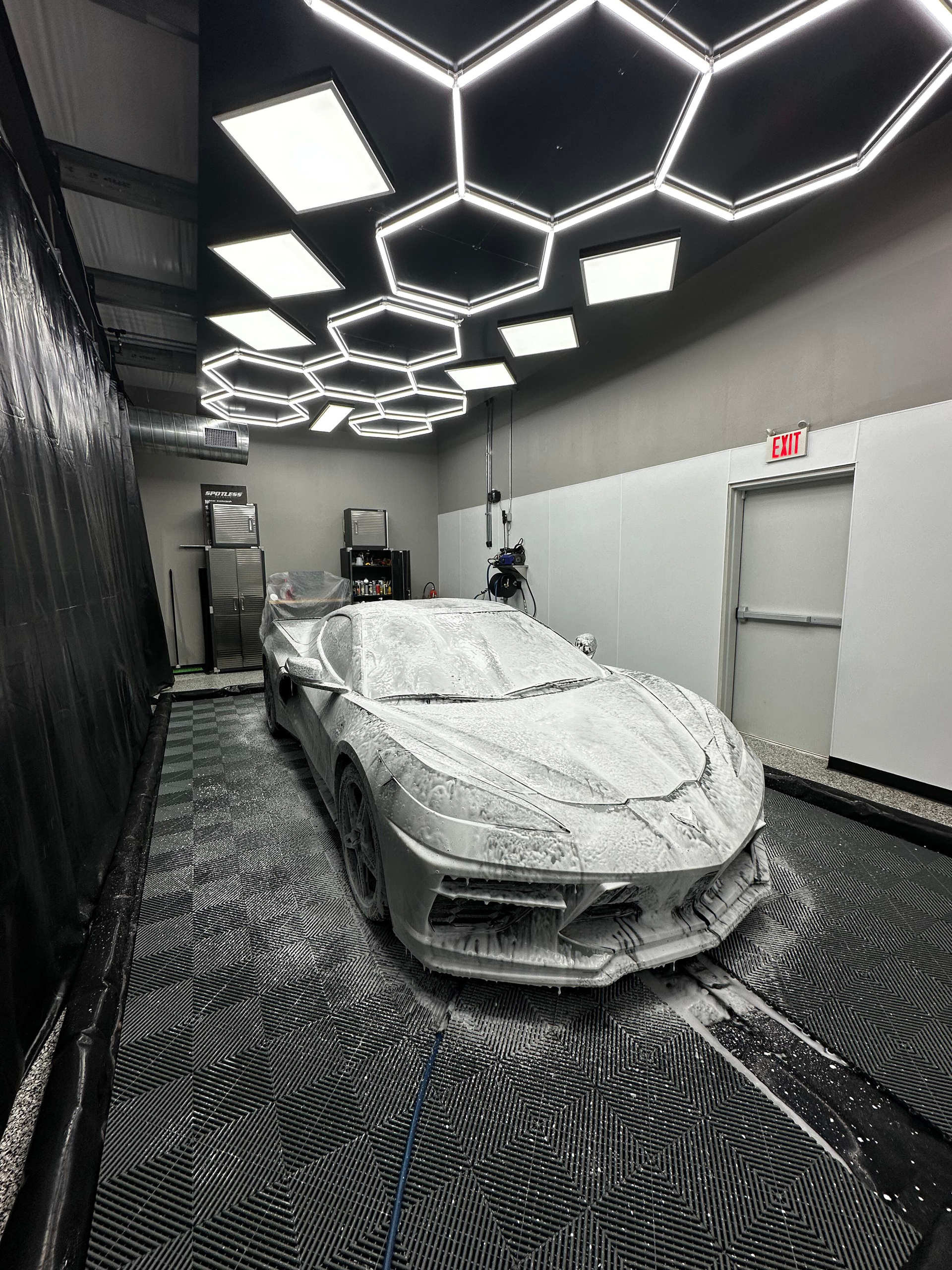 A sports car covered in white foam sits inside a car wash bay with honeycomb ceiling lights.