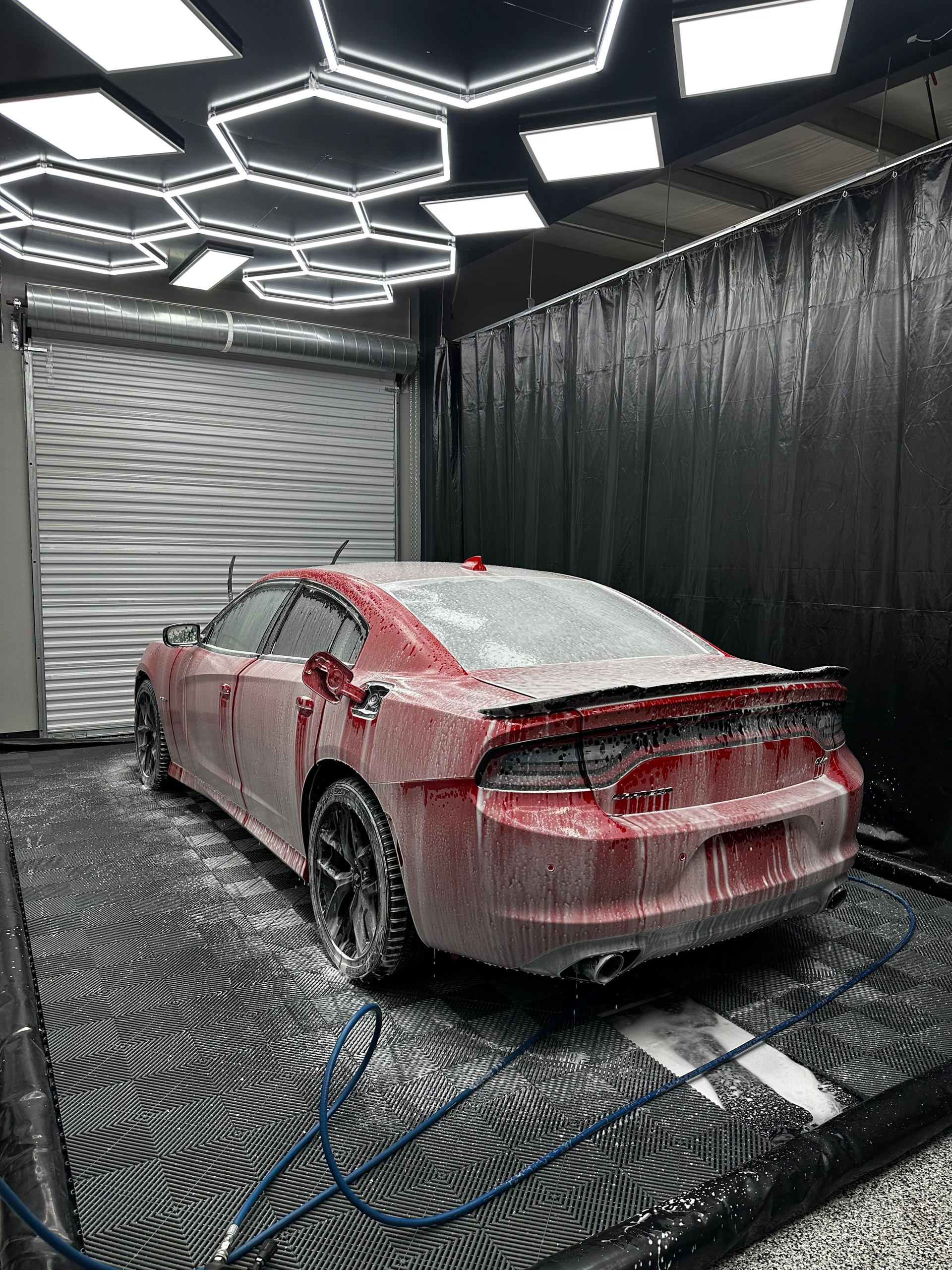Red Dodge Charger being washed in a car wash bay with hexagon-shaped lights overhead. The car is covered in soap.